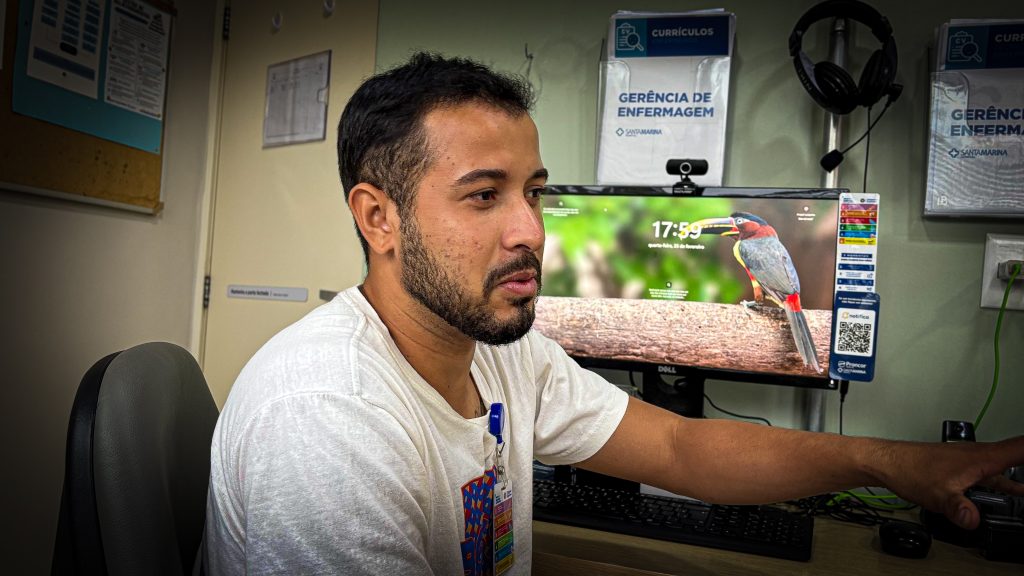 Eurer Eduardo, homem de barba, usando uma camiseta branca, em frente a um computador.
