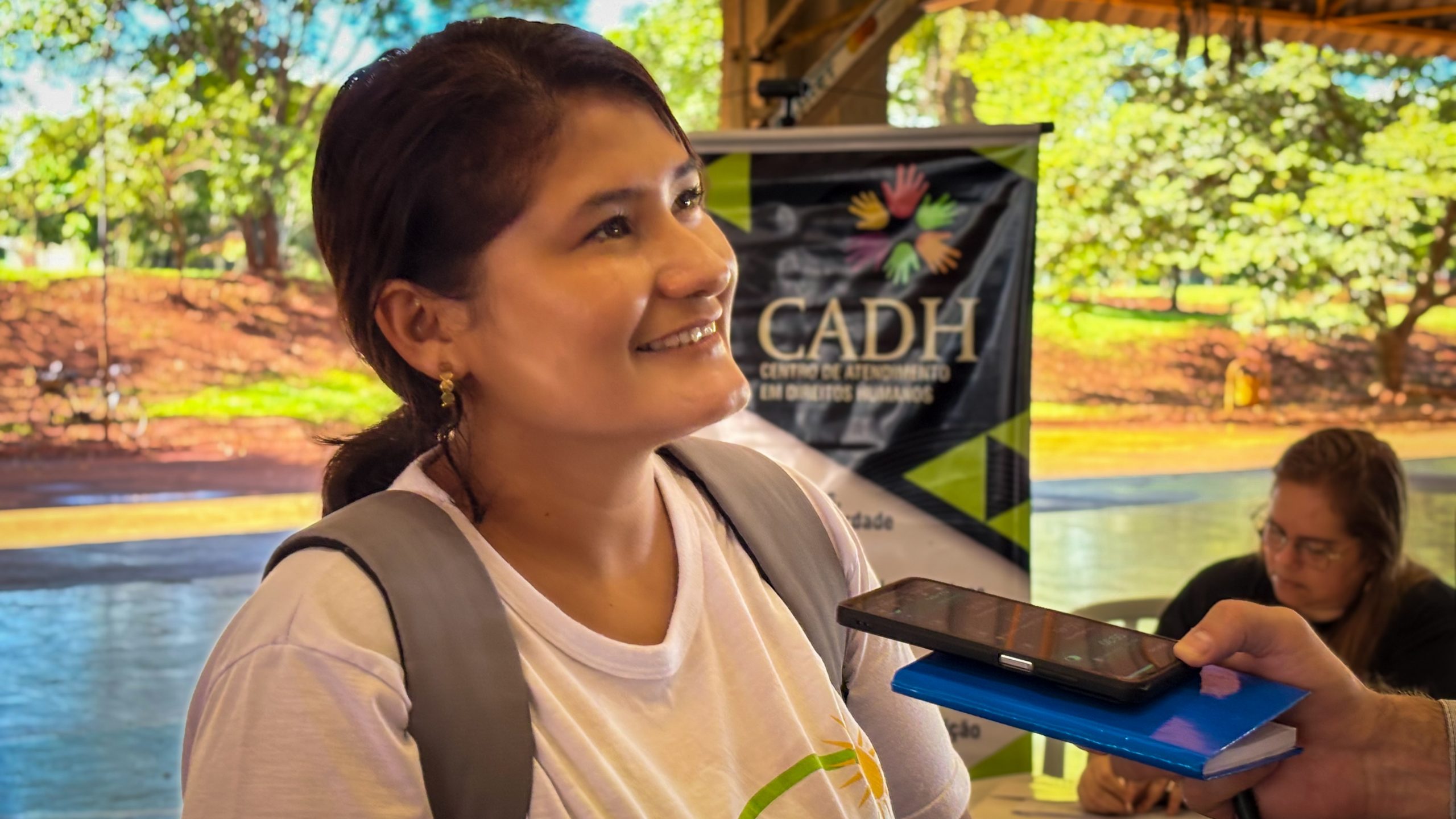 Mulher de camiseta branca e mochila nas costas dá entrevista.