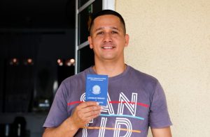 Marcos mostra carteira de trabalho e sorri. Ele tem cabelo curto e usa camiseta.