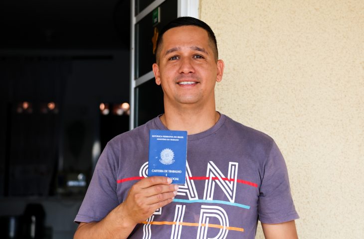 Marcos mostra carteira de trabalho e sorri. Ele tem cabelo curto e usa camiseta.