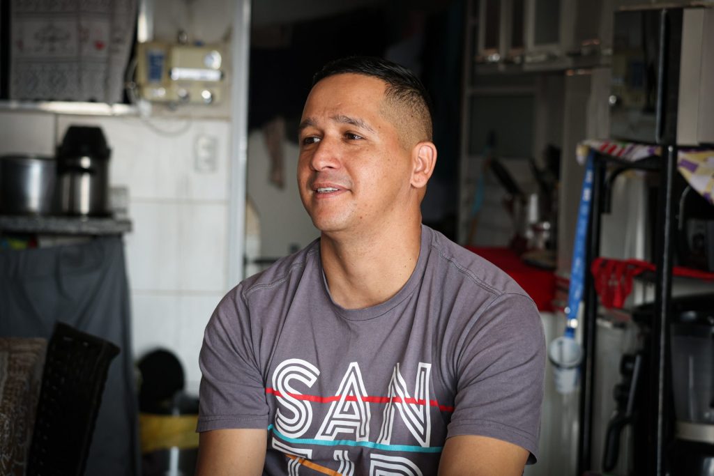Marcos está dentro da casa conversando. Ele é um homem de cabelo curto e usa camiseta cinza.